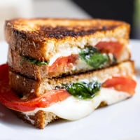 Golden-brown Tomato Basil Grilled Cheese slices are stacked on a rustic wooden board, with melted mozzarella oozing out.  